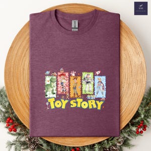 Puede incluir: Una camiseta granate con un gr&aacute;fico colorido de personajes de la pel&iacute;cula animada "Toy Story". El dise&ntilde;o incluye im&aacute;genes de un dinosaurio, Woody, Buzz Lightyear y otros personajes, con las palabras "Toy Story" en amarillo.