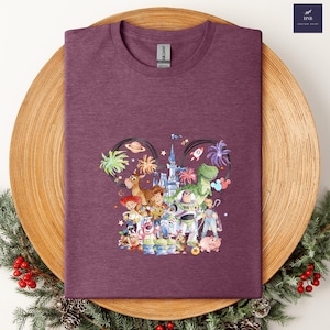 May include: A maroon t-shirt featuring a colorful graphic of Disney's Toy Story characters arranged in a Mickey Mouse ear shape. The design includes Woody, Buzz Lightyear, and other characters. The shirt has a crew neck and a Gildan tag.