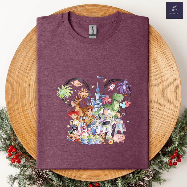 May include: A maroon t-shirt featuring a colorful graphic of Disney's Toy Story characters arranged in a Mickey Mouse ear shape. The design includes Woody, Buzz Lightyear, and other characters. The shirt has a crew neck and a Gildan tag.