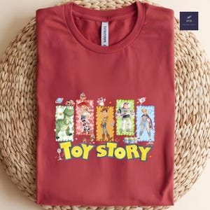 Puede incluir: Una camiseta granate con un gr&aacute;fico colorido de personajes de la pel&iacute;cula animada "Toy Story". El dise&ntilde;o incluye im&aacute;genes de un dinosaurio, Woody, Buzz Lightyear y otros personajes, con las palabras "Toy Story" en amarillo.
