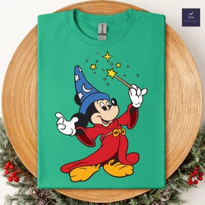 Puede incluir: Una camiseta verde con una imagen de dibujos animados de Mickey Mouse como mago. Mickey lleva un sombrero azul con estrellas blancas, una t&uacute;nica roja y zapatos amarillos. Sostiene una varita m&aacute;gica con estrellas.