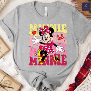 May include: A gray t-shirt featuring a colorful graphic of Minnie Mouse. Minnie is depicted in a pink polka-dot dress and bow, with the word "MINNIE" in a retro font. The shirt has a crew neck and short sleeves.