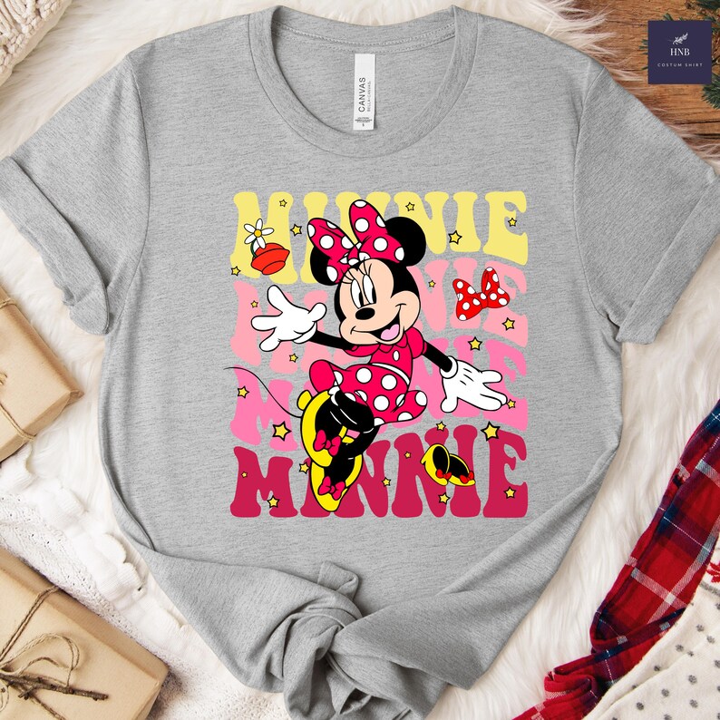 May include: A gray t-shirt featuring a colorful graphic of Minnie Mouse. Minnie is depicted in a pink polka-dot dress and bow, with the word "MINNIE" in a retro font. The shirt has a crew neck and short sleeves.