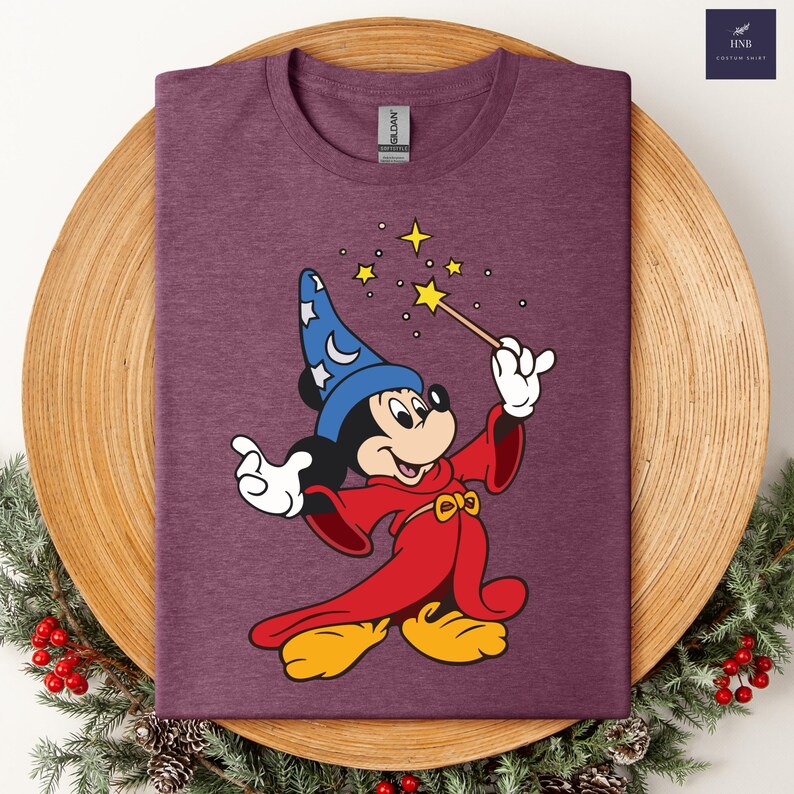 Puede incluir: Camiseta granate con una ilustraci&oacute;n de dibujos animados de Mickey Mouse vestido de mago. Mickey lleva un sombrero de mago azul con estrellas y una t&uacute;nica roja, sosteniendo una varita con estrellas. La camiseta tiene cuello redondo.