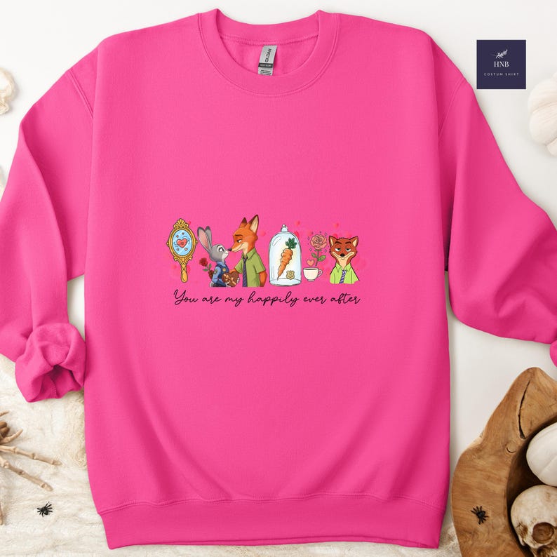 May include: A bright pink sweatshirt featuring a cartoon illustration with the text "You are my happily ever after." The design includes a mirror, a rabbit, a fox, a carrot in a glass dome, and a teacup with a rose.