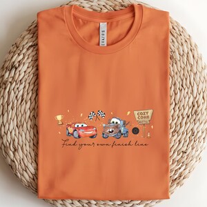 May include: Orange t-shirt featuring a cartoon illustration of two race cars, a trophy, checkered flags, and the text "Find your own finish line." The design includes a sign that says "Cozy Cone Motel."