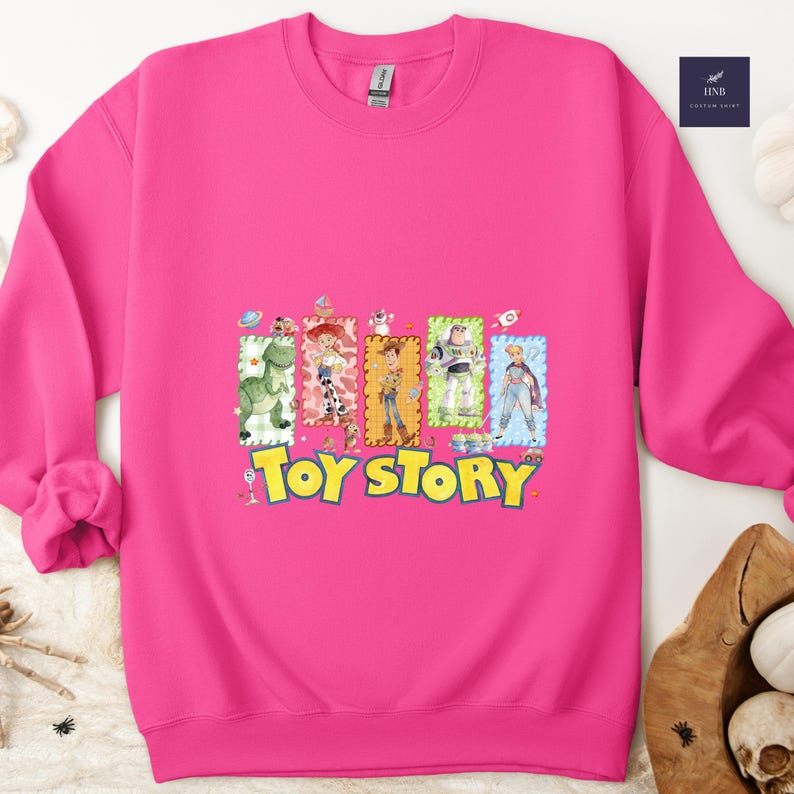 Puede incluir: Una sudadera rosa brillante con un gr&aacute;fico colorido de personajes de la pel&iacute;cula "Toy Story". El gr&aacute;fico incluye im&aacute;genes de Woody, Buzz Lightyear y otros personajes. Las palabras "Toy Story" est&aacute;n impresas en amarillo.
