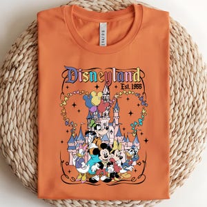 Może przedstawiać: Pomarańczowy t-shirt z okrągłym dekoltem i nadrukiem przedstawiającym zamek Disneyland, Myszkę Miki, Myszkę Minnie, Goofy'ego, Kaczora Donalda i Daisy. Napis głosi Disneyland Est. 1955, w otoczeniu kolorowych balon&oacute;w w kształcie Miki i gwiazdek.