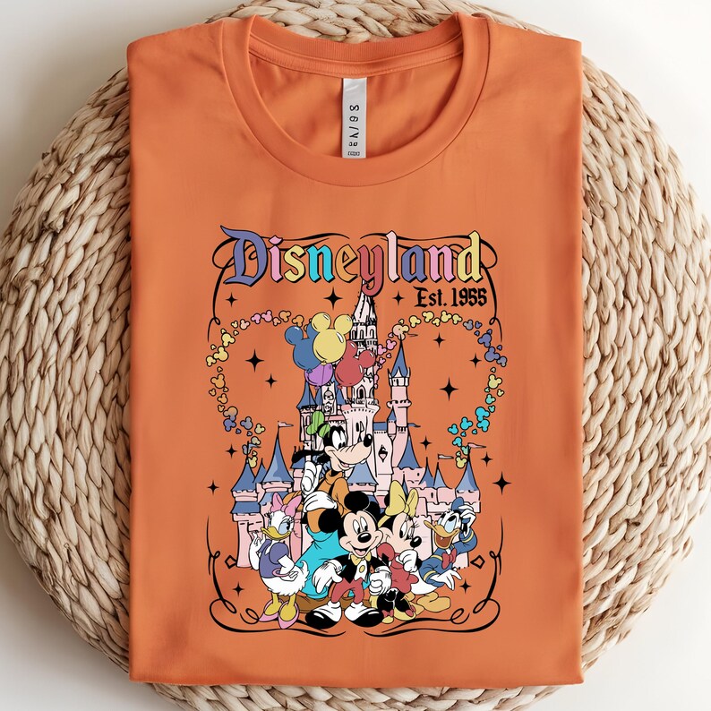 Może przedstawiać: Pomarańczowy t-shirt z okrągłym dekoltem i nadrukiem przedstawiającym zamek Disneyland, Myszkę Miki, Myszkę Minnie, Goofy'ego, Kaczora Donalda i Daisy. Napis głosi Disneyland Est. 1955, w otoczeniu kolorowych balon&oacute;w w kształcie Miki i gwiazdek.