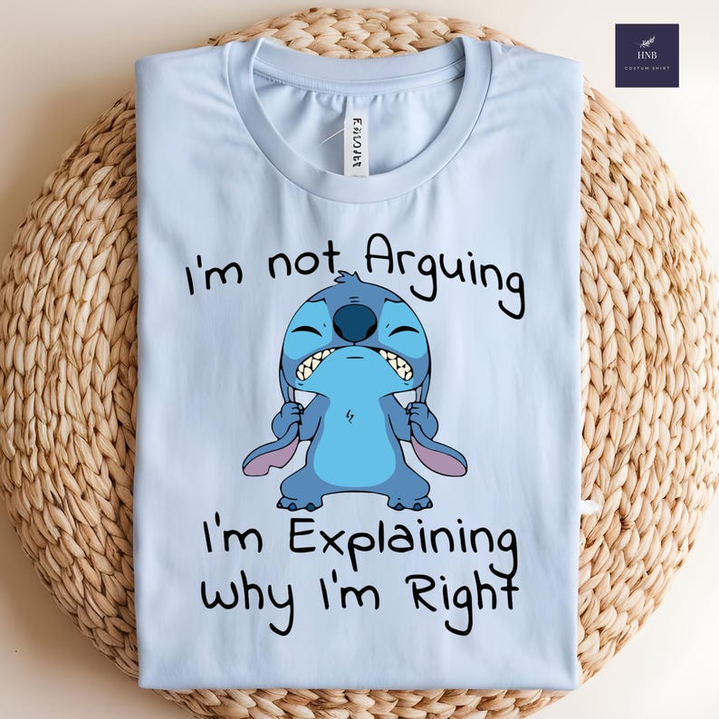 May include: Light blue t-shirt featuring a cartoon character, Stitch, with the text "I'm not Arguing, I'm Explaining why I'm Right." The design is in black and blue. The shirt is folded on a woven surface.