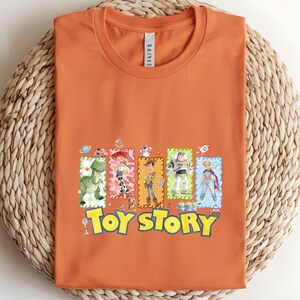 Puede incluir: Camiseta naranja con las palabras "Toy Story" en amarillo y azul, e ilustraciones de personajes de dibujos animados. La camiseta tiene cuello redondo y est&aacute; doblada cuidadosamente.
