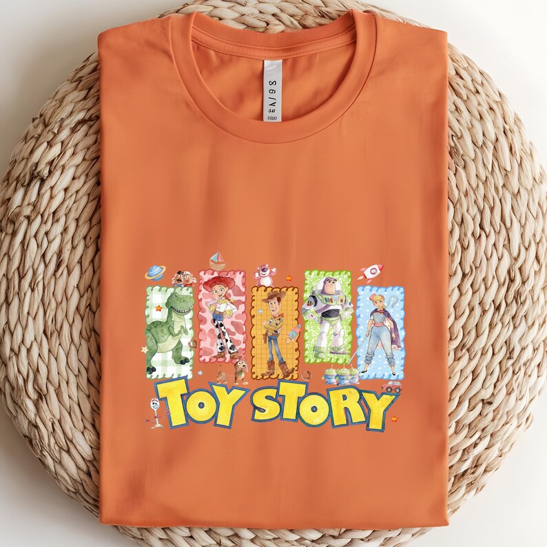 Puede incluir: Camiseta naranja con las palabras "Toy Story" en amarillo y azul, e ilustraciones de personajes de dibujos animados. La camiseta tiene cuello redondo y est&aacute; doblada cuidadosamente.