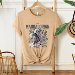 Puede incluir: Camiseta color arena con la frase "THE MANDALORIAN" y un dise&ntilde;o del casco de Mandalorian y Grogu rodeados de flores. El dise&ntilde;o es en tonos grises, morados y verdes. La camiseta est&aacute; colgada en una percha de madera.
