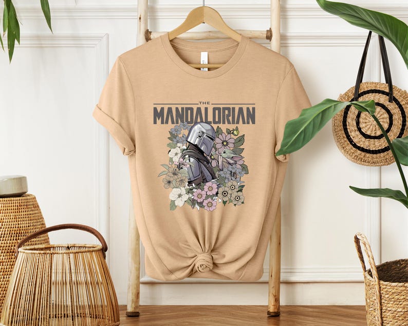 Puede incluir: Camiseta color arena con la frase "THE MANDALORIAN" y un dise&ntilde;o del casco de Mandalorian y Grogu rodeados de flores. El dise&ntilde;o es en tonos grises, morados y verdes. La camiseta est&aacute; colgada en una percha de madera.