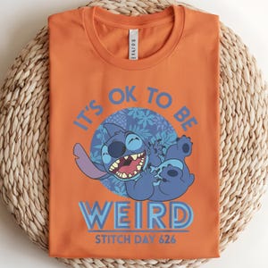 Puede incluir: Camiseta naranja con un dise&ntilde;o de Stitch de Lilo & Stitch. El dise&ntilde;o incluye el texto "IT'S OK TO BE WEIRD" y "STITCH DAY 626". El personaje de dibujos animados Stitch es azul y sonr&iacute;e.