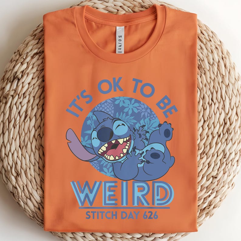 Puede incluir: Camiseta naranja con un dise&ntilde;o de Stitch de Lilo & Stitch. El dise&ntilde;o incluye el texto "IT'S OK TO BE WEIRD" y "STITCH DAY 626". El personaje de dibujos animados Stitch es azul y sonr&iacute;e.
