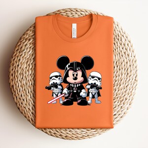 Pu&ograve; includere: T-shirt arancione con un'illustrazione a cartoni animati di Topolino nei panni di Darth Vader, affiancato da due Stormtrooper. Il disegno include una spada laser rossa. La maglietta &egrave; piegata su una superficie tessuta.