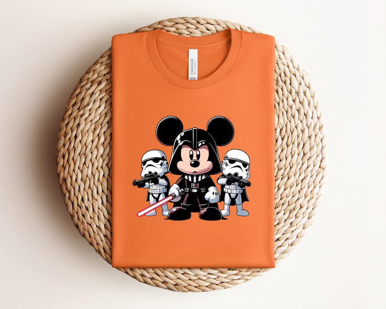 Pu&ograve; includere: T-shirt arancione con un'illustrazione a cartoni animati di Topolino nei panni di Darth Vader, affiancato da due Stormtrooper. Il disegno include una spada laser rossa. La maglietta &egrave; piegata su una superficie tessuta.