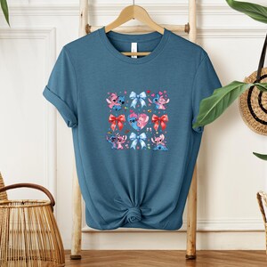 May include: A teal blue t-shirt featuring a colorful graphic of cartoon characters, bows, and hearts. The design includes pink, blue, and red elements. The shirt is displayed on a wooden hanger.