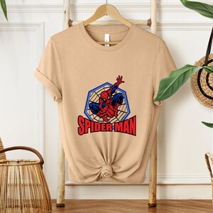 May include: A tan t-shirt featuring a graphic of Spider-Man in a blue and red costume, within a blue web-like hexagon. The word "SPIDER-MAN" is printed in red below the image. The shirt is displayed on a wooden hanger.