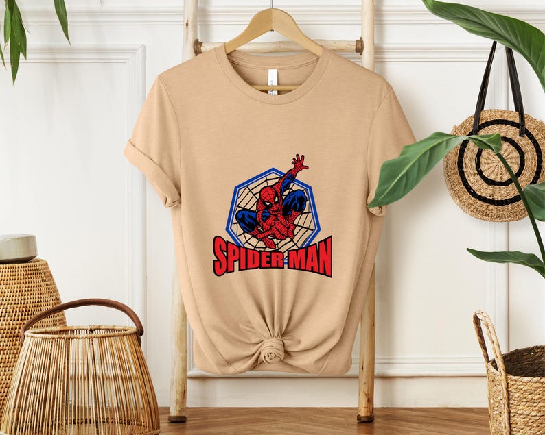 May include: A tan t-shirt featuring a graphic of Spider-Man in a blue and red costume, within a blue web-like hexagon. The word "SPIDER-MAN" is printed in red below the image. The shirt is displayed on a wooden hanger.