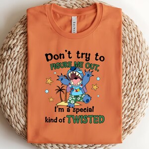 May include: Orange t-shirt featuring a cartoon character with the text "Don't try to FIGURE ME OUT, I'm a special kind of TWISTED." The character is blue with large eyes and an open mouth, wearing a Hawaiian shirt.