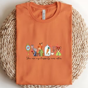 May include: Orange t-shirt featuring a cartoon design with characters and the text "You are my happily ever after." The design includes a mirror, a rabbit, a fox, a carrot in a glass dome, a rose, and a teacup.