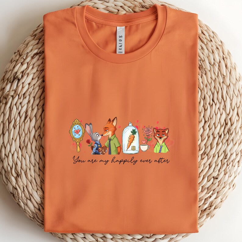 May include: Orange t-shirt featuring a cartoon design with characters and the text "You are my happily ever after." The design includes a mirror, a rabbit, a fox, a carrot in a glass dome, a rose, and a teacup.