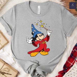 Puede incluir: Camiseta gris con una imagen de dibujos animados de Mickey Mouse como mago. Mickey lleva un sombrero de mago azul con lunas y estrellas, una t&uacute;nica roja y zapatos amarillos. Sostiene una varita con estrellas.
