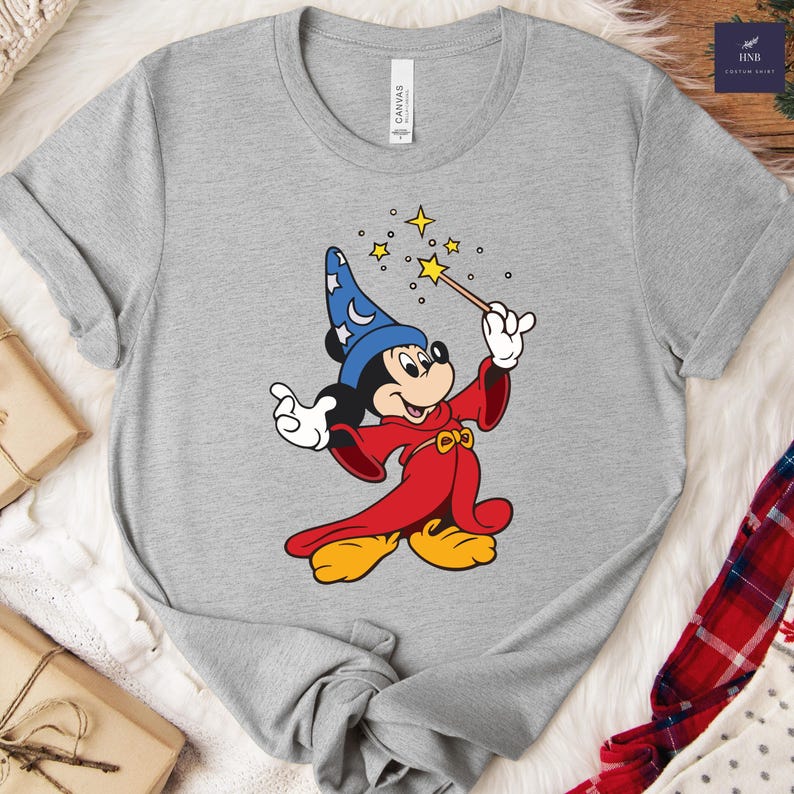 Puede incluir: Camiseta gris con una imagen de dibujos animados de Mickey Mouse como mago. Mickey lleva un sombrero de mago azul con lunas y estrellas, una t&uacute;nica roja y zapatos amarillos. Sostiene una varita con estrellas.