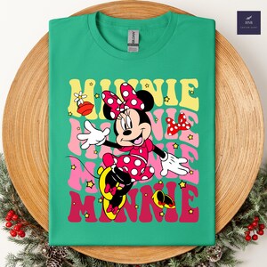 May include: Green t-shirt featuring Minnie Mouse in a red polka dot dress and bow. The word "MINNIE" is printed in layered retro font with stars. A fun and colorful design for Disney fans.