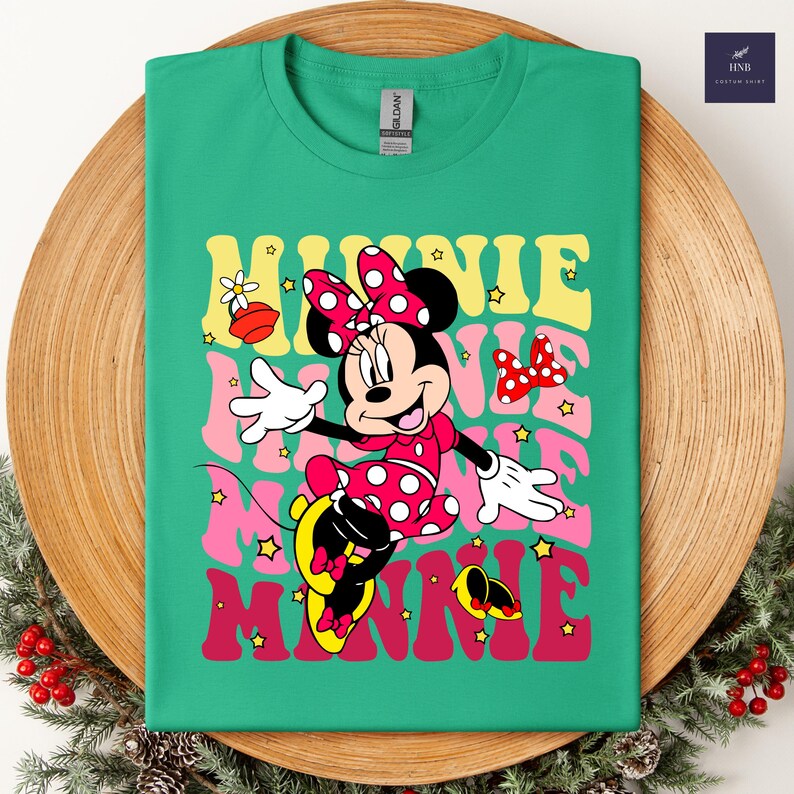 May include: Green t-shirt featuring Minnie Mouse in a red polka dot dress and bow. The word "MINNIE" is printed in layered retro font with stars. A fun and colorful design for Disney fans.