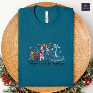K&ouml;nnte beinhalten: T&uuml;rkisfarbenes T-Shirt mit einer Cartoon-Illustration von Tierfiguren. Das Design umfasst einen Biber, einen Fuchs, ein Kaninchen und eine Schlange, mit dem Text "Anyone can be anything". Das T-Shirt ist aus Canvas-Material.