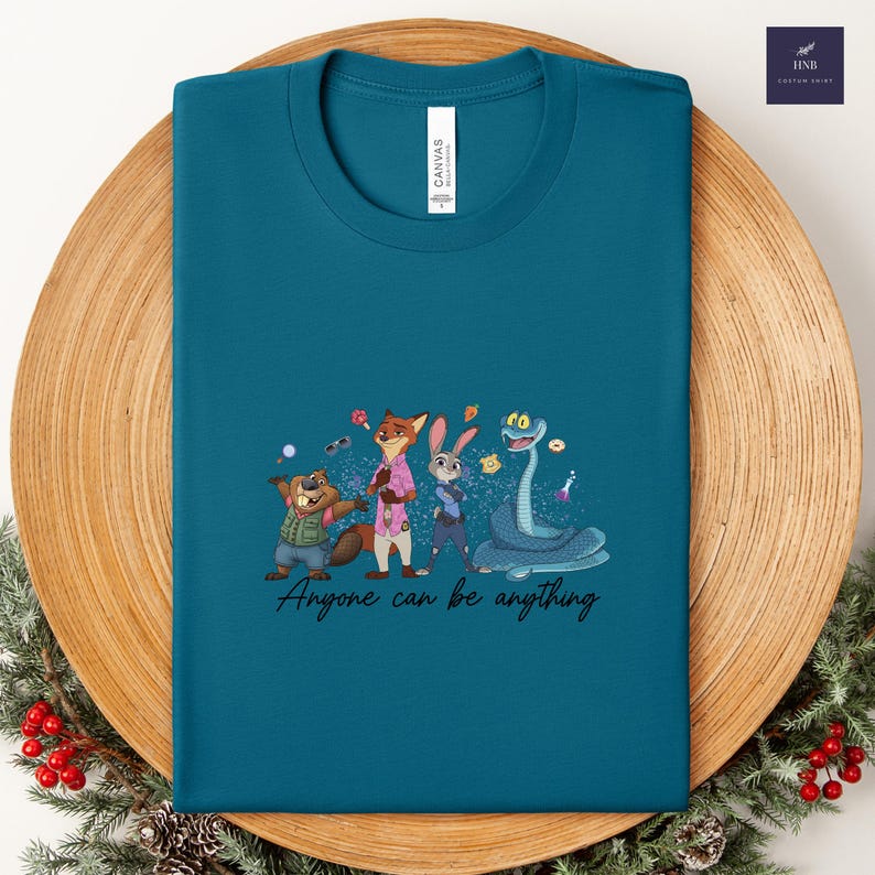 K&ouml;nnte beinhalten: T&uuml;rkisfarbenes T-Shirt mit einer Cartoon-Illustration von Tierfiguren. Das Design umfasst einen Biber, einen Fuchs, ein Kaninchen und eine Schlange, mit dem Text "Anyone can be anything". Das T-Shirt ist aus Canvas-Material.