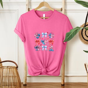 May include: A bright pink t-shirt featuring a colorful graphic of cartoon characters with bows. The graphic includes blue, pink, and red bows, and the characters are in various poses. The shirt is hanging on a wooden hanger.