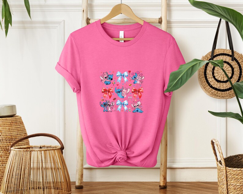 May include: A bright pink t-shirt featuring a colorful graphic of cartoon characters with bows. The graphic includes blue, pink, and red bows, and the characters are in various poses. The shirt is hanging on a wooden hanger.