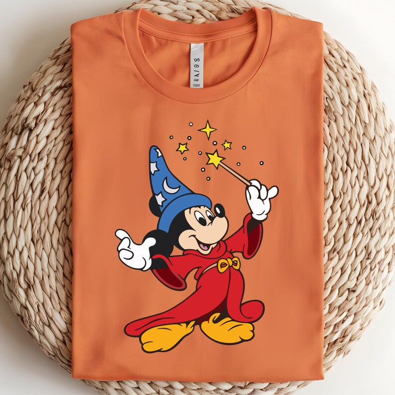 Puede incluir: Camiseta naranja con una ilustraci&oacute;n de dibujos animados de Mickey Mouse como mago. Mickey lleva un sombrero de mago azul con estrellas blancas, una t&uacute;nica roja, zapatos amarillos y guantes blancos. Sostiene una varita con estrellas. La camiseta est&aacute; doblada.