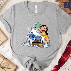 Pode incluir: Uma camiseta cinza com uma ilustra&ccedil;&atilde;o de desenho animado de Lilo e Stitch. Lilo est&aacute; despejando sab&atilde;o em Stitch, que est&aacute; em uma banheira de madeira. A camiseta tem gola redonda e mangas curtas. A marca "Canvas" est&aacute; impressa na camiseta.