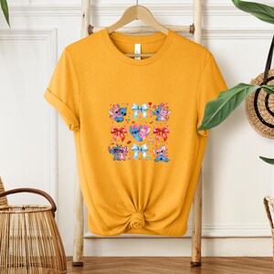 May include: Mustard yellow t-shirt featuring a colorful graphic of cartoon characters and bows. The graphic includes blue, pink, and red elements. The shirt is hanging on a wooden hanger against a white wall.