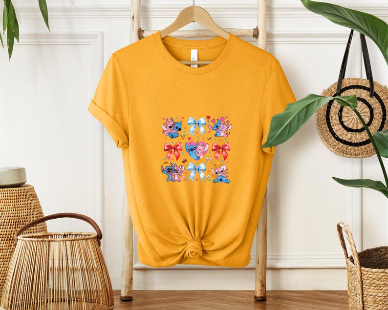 May include: Mustard yellow t-shirt featuring a colorful graphic of cartoon characters and bows. The graphic includes blue, pink, and red elements. The shirt is hanging on a wooden hanger against a white wall.