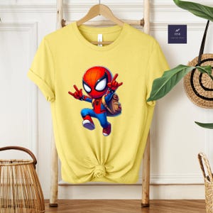 Puede incluir: Camiseta amarilla con un dise&ntilde;o de Spider-Man de dibujos animados en rojo y azul, con mochila y pose din&aacute;mica. La camiseta est&aacute; colgada en un perchero de madera. Un peque&ntilde;o cuadrado azul con el texto "HNB CUSTOM SHIRT" es visible.