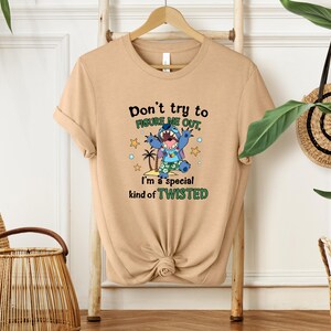 May include: A tan t-shirt with the text "Don't try to figure me out, I'm a special kind of TWISTED" and a cartoon character graphic. The graphic features a blue alien-like creature with a tropical scene. The shirt is displayed on a wooden hanger.