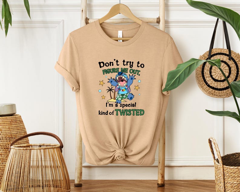 May include: A tan t-shirt with the text "Don't try to figure me out, I'm a special kind of TWISTED" and a cartoon character graphic. The graphic features a blue alien-like creature with a tropical scene. The shirt is displayed on a wooden hanger.