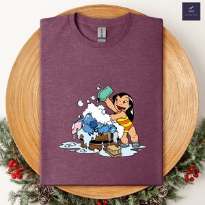 Pode incluir: Camiseta bord&ocirc; com um gr&aacute;fico de Lilo lavando Stitch em uma banheira de madeira com bolhas. Lilo est&aacute; usando um mai&ocirc; listrado amarelo e laranja. A camisa tem gola redonda e uma etiqueta Gildan.