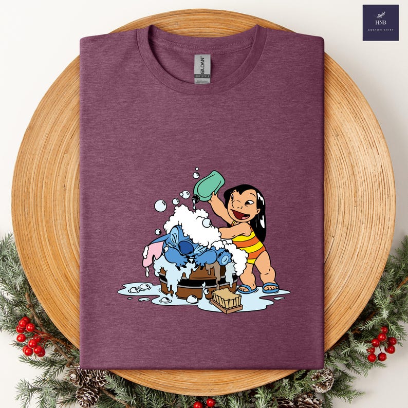 Pode incluir: Camiseta bord&ocirc; com um gr&aacute;fico de Lilo lavando Stitch em uma banheira de madeira com bolhas. Lilo est&aacute; usando um mai&ocirc; listrado amarelo e laranja. A camisa tem gola redonda e uma etiqueta Gildan.