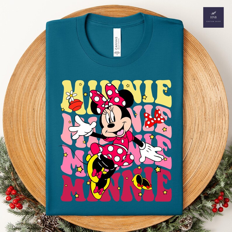May include: Teal t-shirt featuring a colorful graphic of Minnie Mouse. The design includes the word "MINNIE" in retro-style lettering, along with Minnie in a polka-dot dress and bow. The shirt is displayed on a wooden tray.