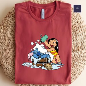 Pode incluir: Uma camiseta bord&ocirc; com uma ilustra&ccedil;&atilde;o de desenho animado de Lilo lavando Stitch em uma tina de madeira. Lilo est&aacute; usando um mai&ocirc; listrado amarelo e laranja. A camisa tem gola redonda e mangas curtas.