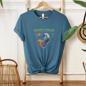 May include: A teal t-shirt with a graphic of the Mandalorian and Grogu. The word "MANDALORIAN" is printed above the image in a colourful, retro font. The design includes a triangle, a sun, and stars.