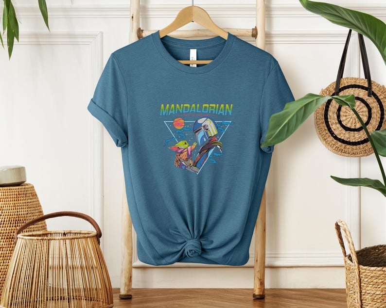 May include: A teal t-shirt with a graphic of the Mandalorian and Grogu. The word "MANDALORIAN" is printed above the image in a colourful, retro font. The design includes a triangle, a sun, and stars.
