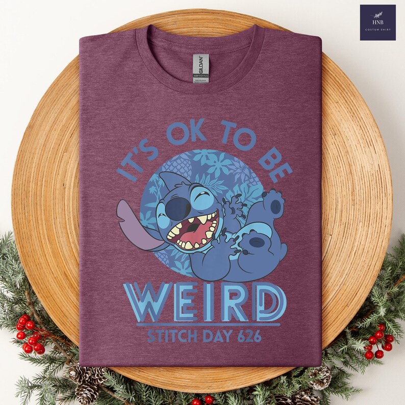 Puede incluir: Camiseta granate con un dise&ntilde;o del personaje Stitch de Lilo & Stitch. El dise&ntilde;o incluye el texto "IT'S OK TO BE WEIRD" y "STITCH DAY 626". El dibujo animado de Stitch es azul y se r&iacute;e.
